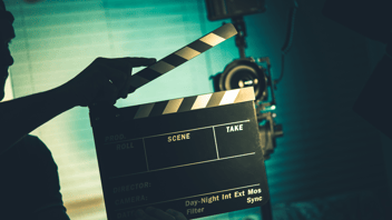 On set with a slate, a video camera is seen in the background.